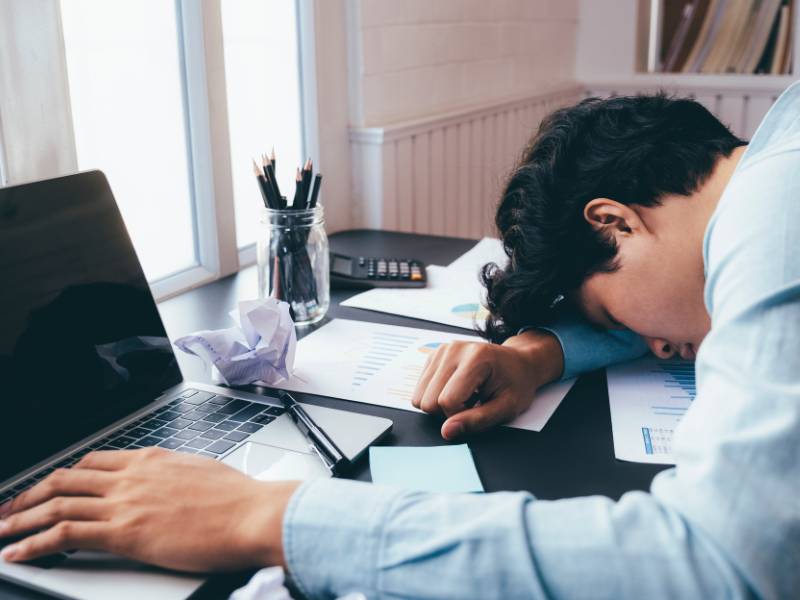 パソコン作業に疲れて机の上に突っ伏して寝ている会社員男性の画像